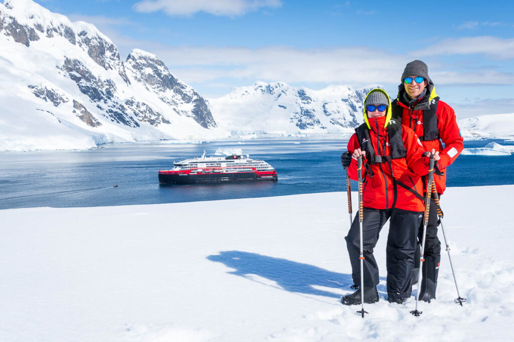 Seb and Ieva standing on the snow with Salpêtrière Bay in the background with the Hurtigruten expedition ship.
