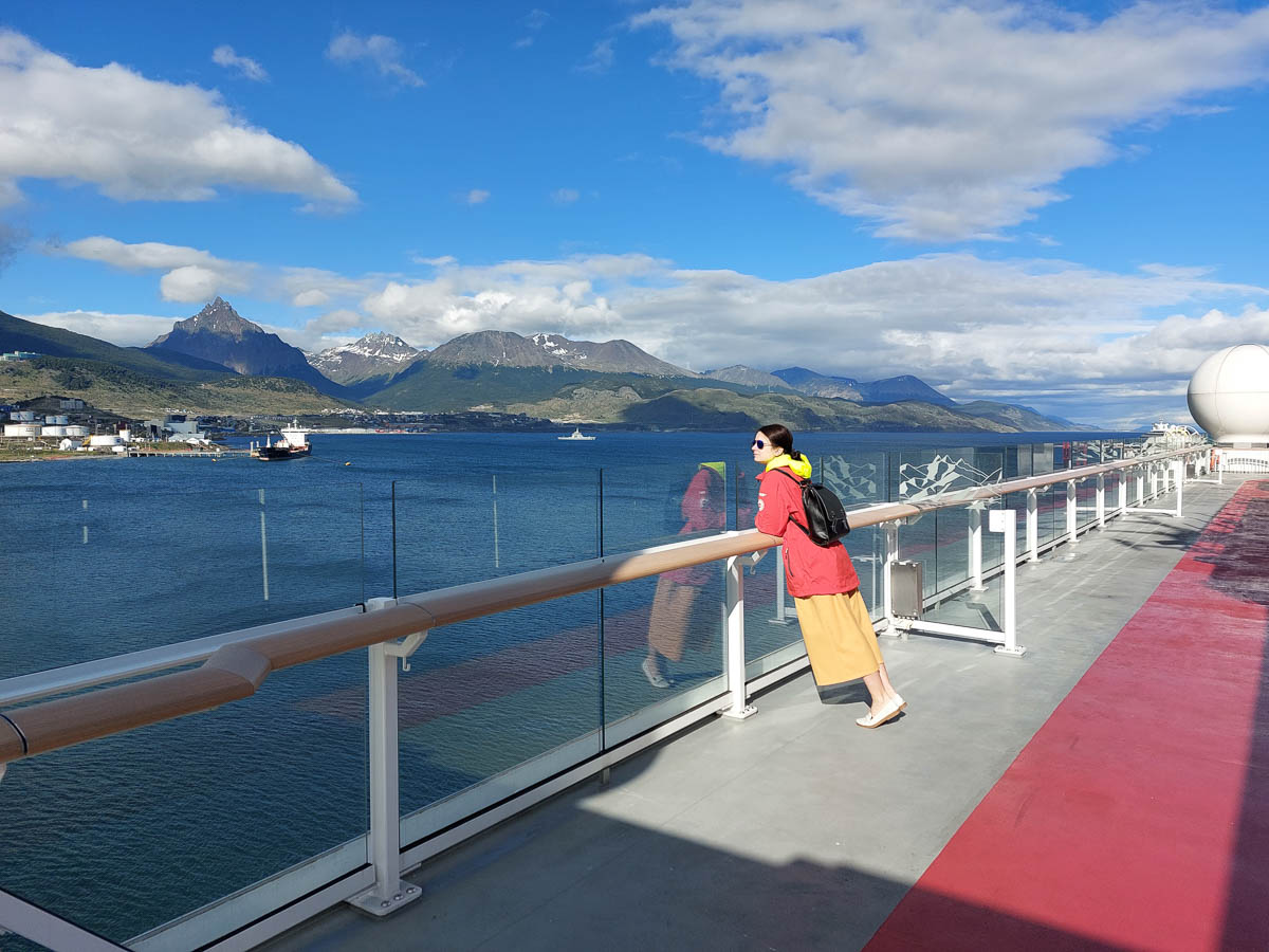 A woman in red jacket and yellow trousers on deck of a cruise ship looking at the scenery of Ushuaia.