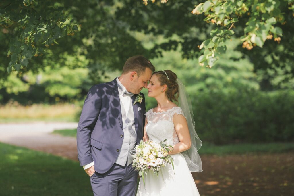 Bride and groom embracing under trees in a sunlit garden, sharing a tender moment during their wedding day.