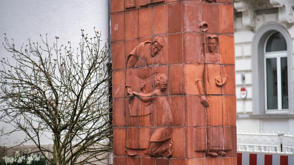 Terracotta sculpture depicting human figures on a building façade next to a leafless tree.
