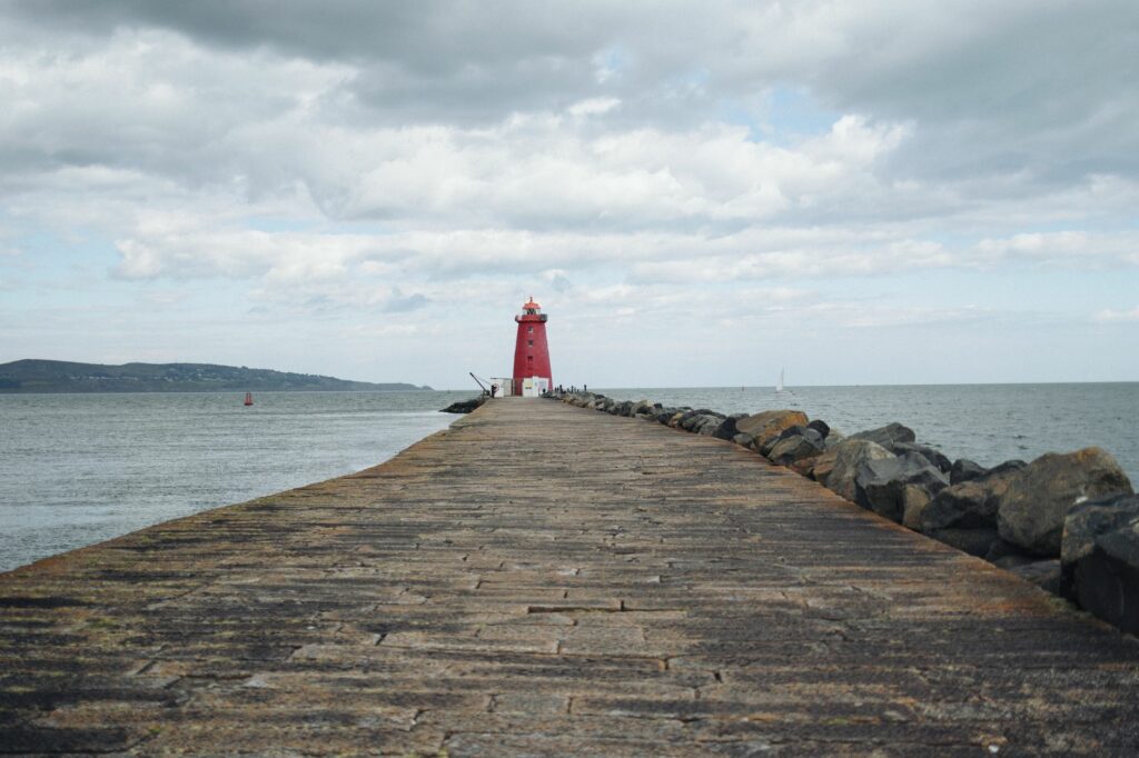 A red lighthouse stands at the end of a rocky pier under a cloudy sky by the sea.