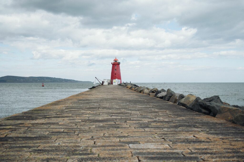 Scenic view of a red lighthouse on a long pier extending into the ocean under cloudy skies.