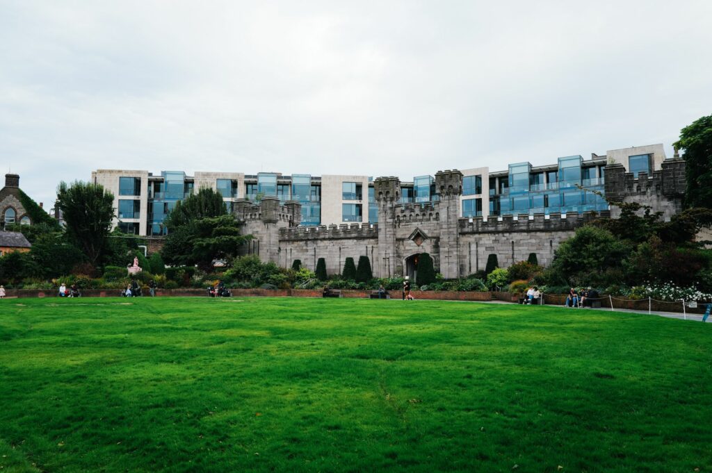 A historic castle wall with a modern building and green lawn in the foreground