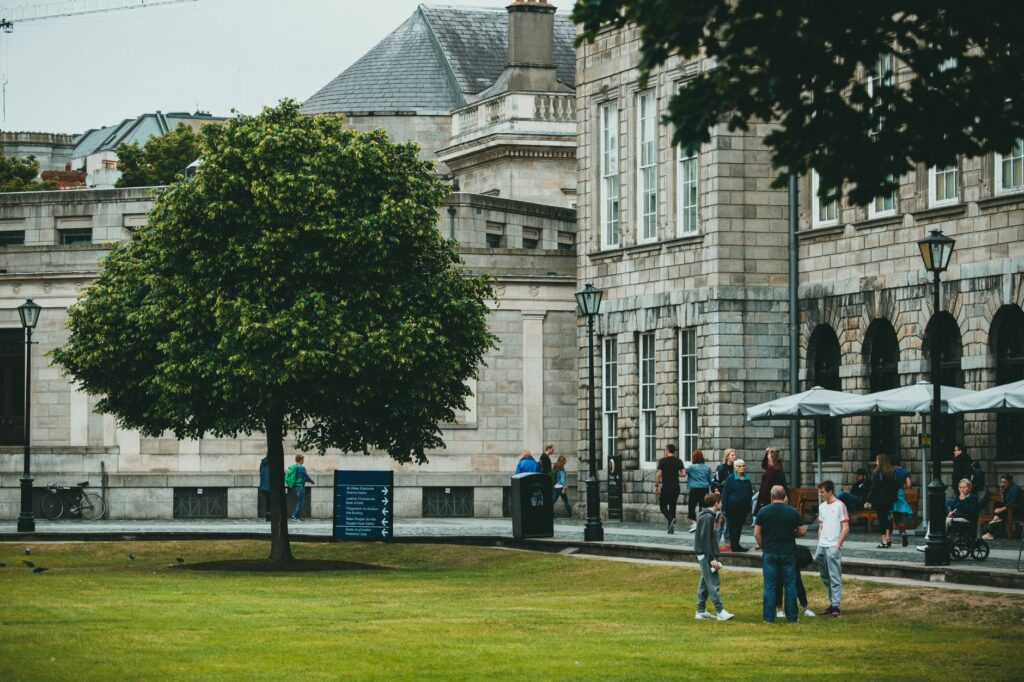 People gathering near a historical building in an urban park, surrounded by greenery.