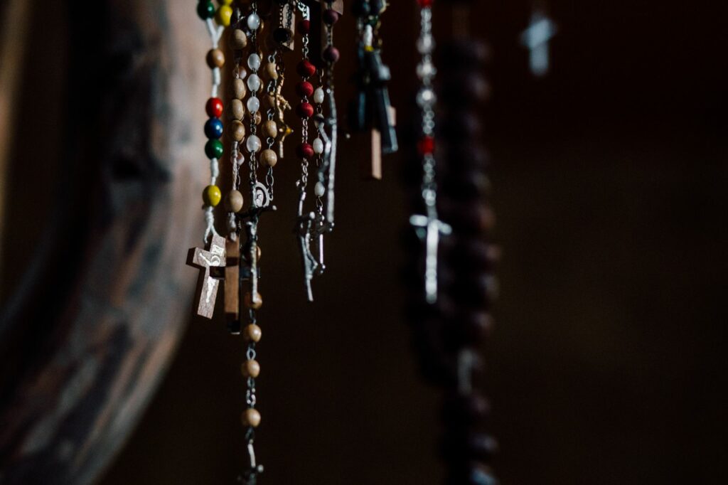 A collection of colorful rosary beads with crosses hanging in a dark indoor setting.
