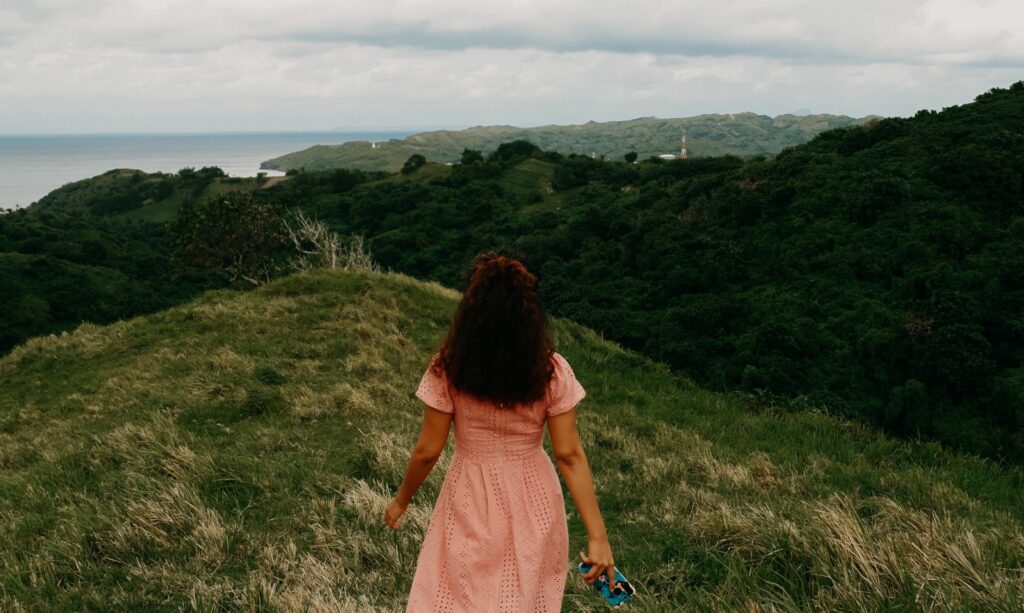 A woman in a pink dress walks towards the scenic hills of Batanes, Philippines.