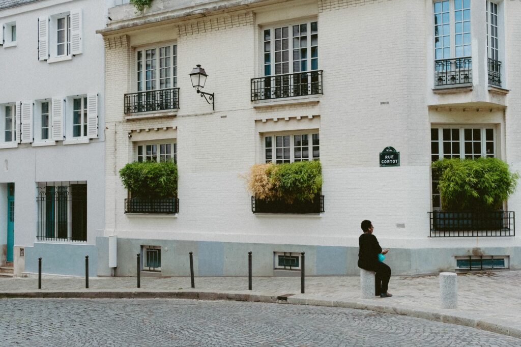 Southern European street scene with architecture, plants, and a person sitting on the sidewalk.