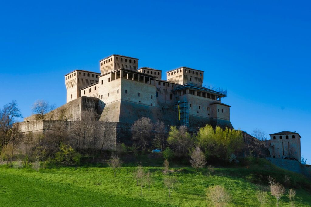 A grand medieval castle sits atop a hill against a clear blue sky, surrounded by lush greenery.