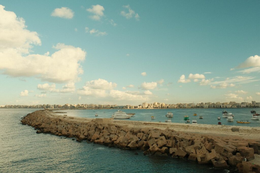 A picturesque view of a Mediterranean coastline with boats and distant city skyline under a partly cloudy sky.