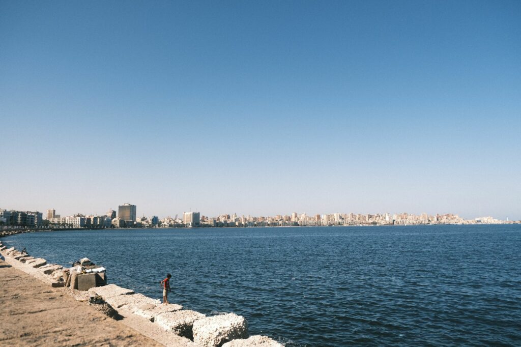 Serene view of Alexandria's coastline with cityscape in the background under a clear sky.