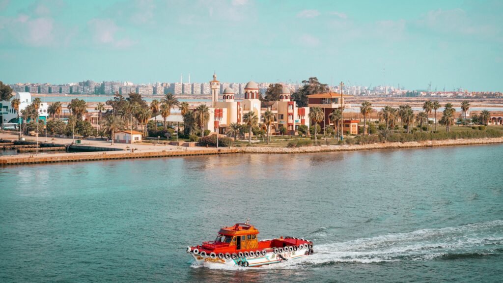 A scenic view of a red boat cruising near a city with palm trees and vibrant architecture.