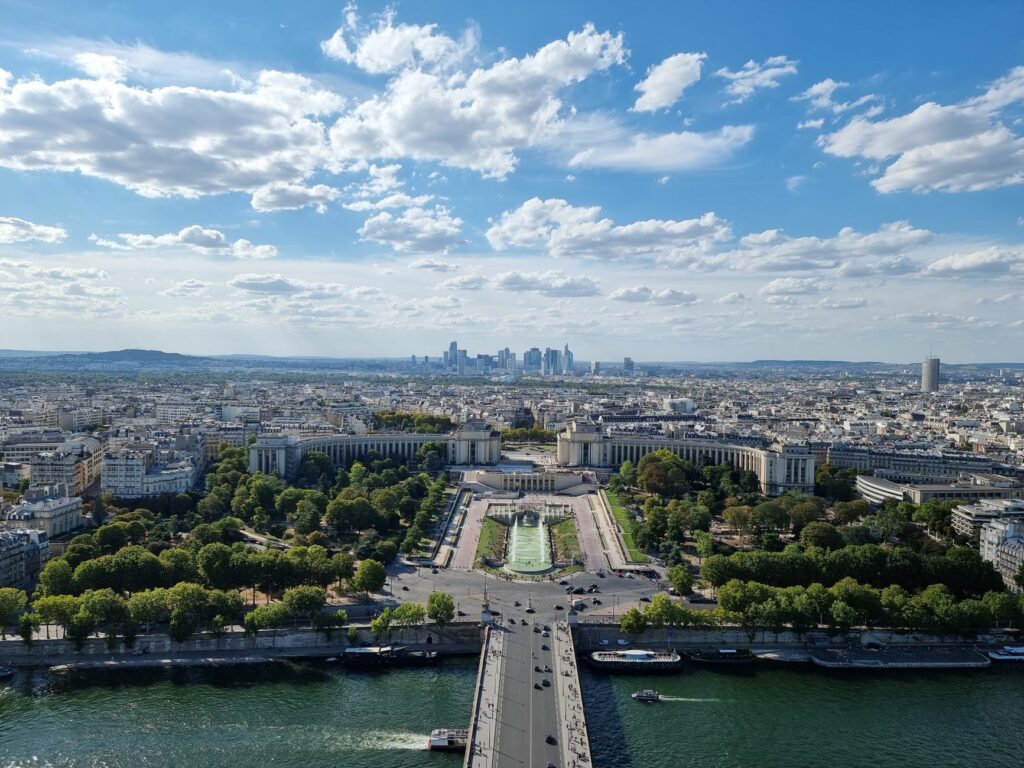 Aerial view of Paris featuring Trocadéro Gardens and La Défense skyline under a bright sky.